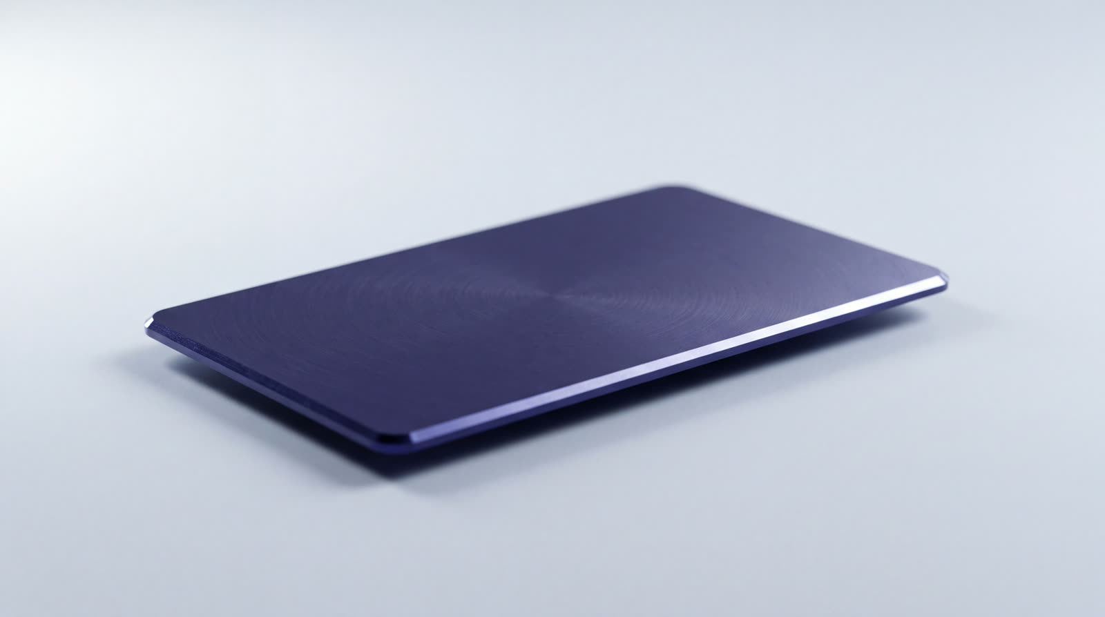 A single thin machined card of indigo anodised metal lying on a pale ground, its face blank and one milled edge catching the key light.